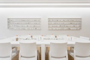 White chairs surround a white table with glue, white rolls of string and broken ceramic pieces atop it.